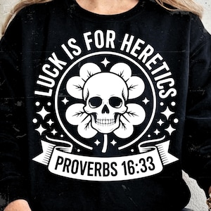 Peut inclure: Sweat-shirt noir avec un motif blanc. Le motif représente un crâne à l'intérieur d'une fleur, entouré du texte "LUCK IS FOR HERETICS" et "PROVERBS 16:33". Le design comprend des étoiles et des rubans.