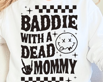 Può includere: Felpa bianca con la scritta "BADIE WITH A DEAD MOMMY" in nero. Il design include una faccina sorridente con gli occhi sbarrati e un gesto della mano. La parte superiore e inferiore del design hanno un motivo a scacchi.