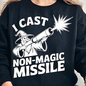 Könnte beinhalten: Schwarzes Sweatshirt mit weißer Grafik eines Zauberers, der eine Kanone abfeuert, mit dem Text "I CAST NON-MAGIC MISSILE". Der Zauberer trägt Hut und Robe. Das Design beinhaltet einen Lichtblitz aus der Kanone.