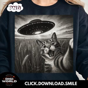 May include: Black and white graphic of a cat taking a selfie with a UFO hovering above a wheat field. The cat has wide eyes and an open mouth. The image is on a black sweatshirt. Text at the bottom says "CLICK.DOWNLOAD.SMILE."