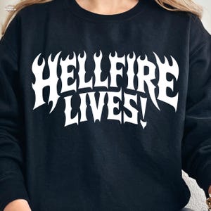 Op de afbeelding: Zwart sweatshirt met de witte tekst "HELLFIRE LIVES!" in een vet, vlamachtig lettertype. Het ontwerp is gecentreerd op de borst. Het sweatshirt is effen zwart van kleur.