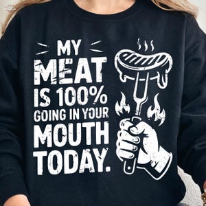 Pode incluir: Camisola preta com um gráfico branco. O gráfico apresenta o texto "MY MEAT IS 100% GOING IN YOUR MOUTH TODAY." com uma ilustração de uma salsicha num garfo, chamas e uma mão segurando o garfo. É uma peça de vestuário casual.