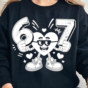 May include: Black sweatshirt featuring a white cartoon heart character wearing glasses and holding the numbers 6 and 7. The design includes hearts, stars, and sneakers, creating a playful and celebratory theme.