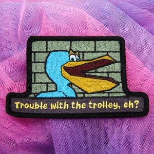 May include: Embroidered patch featuring a cartoon pelican with a blue head and yellow beak, set against a brick wall background. The patch has the text "Trouble with the trolley, eh?" in a brown font on a black border.