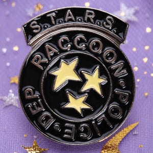 以下が含まれることがあります： 縁に「Raccoon Police Dept.」と「S.T.A.R.S.」の文字が書かれた黒と銀の七宝焼きピン。中央に黄色い星が3つある。