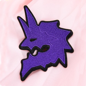 May include: A purple embroidered patch with a black border. The patch features a stylized, jagged design, possibly a creature or symbol. The patch is set against a soft, pink fabric background.
