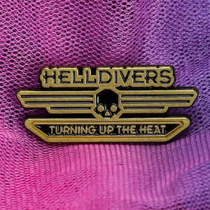 Könnte beinhalten: Goldfarbene Metallanstecknadel mit der Aufschrift "HELLDIVERS" und "TURNING UP THE HEAT". Die Anstecknadel zeigt ein Totenkopf- und gekreuzte Knochen-Design. Der Hintergrund ist ein Farbverlauf aus Rosa und Lila.