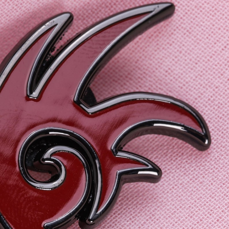 May include: A red and black enamel pin with a flame-like design. The pin features a glossy red center with black outlining and a swirling detail. The pin is set against a pink fabric background.