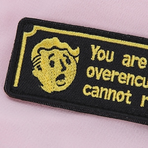 May include: Embroidered black patch with a yellow border and text that reads "You are overencumbered, cannot run." Features a yellow cartoon character's face. The patch is rectangular and has a stitched border.