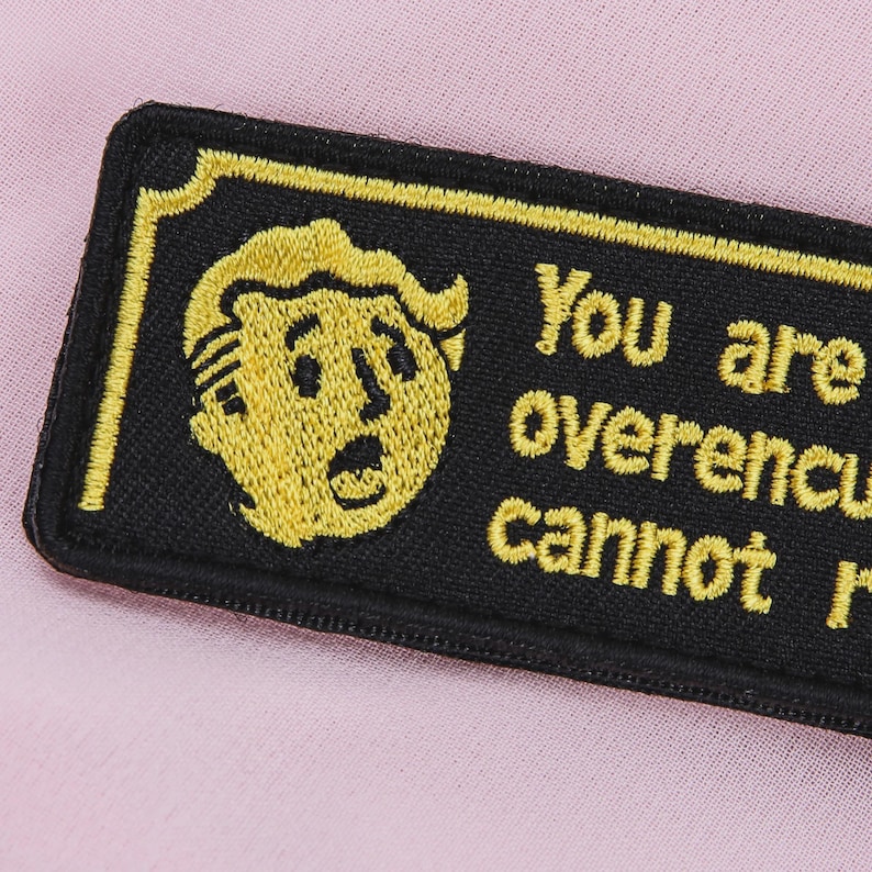 May include: Embroidered black patch with a yellow border and text that reads "You are overencumbered, cannot run." Features a yellow cartoon character's face. The patch is rectangular and has a stitched border.