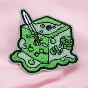 Peut inclure: Patch brodé vert représentant un dé avec une épée qui en sort. Le dé est entouré d'une bordure verte.