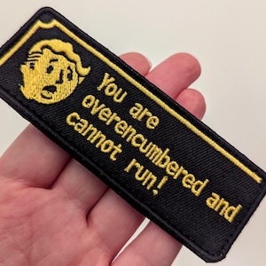 May include: Black rectangular embroidered patch with yellow text and a yellow cartoon character. The text reads "You are overencumbered and cannot run!" The patch has a yellow border.