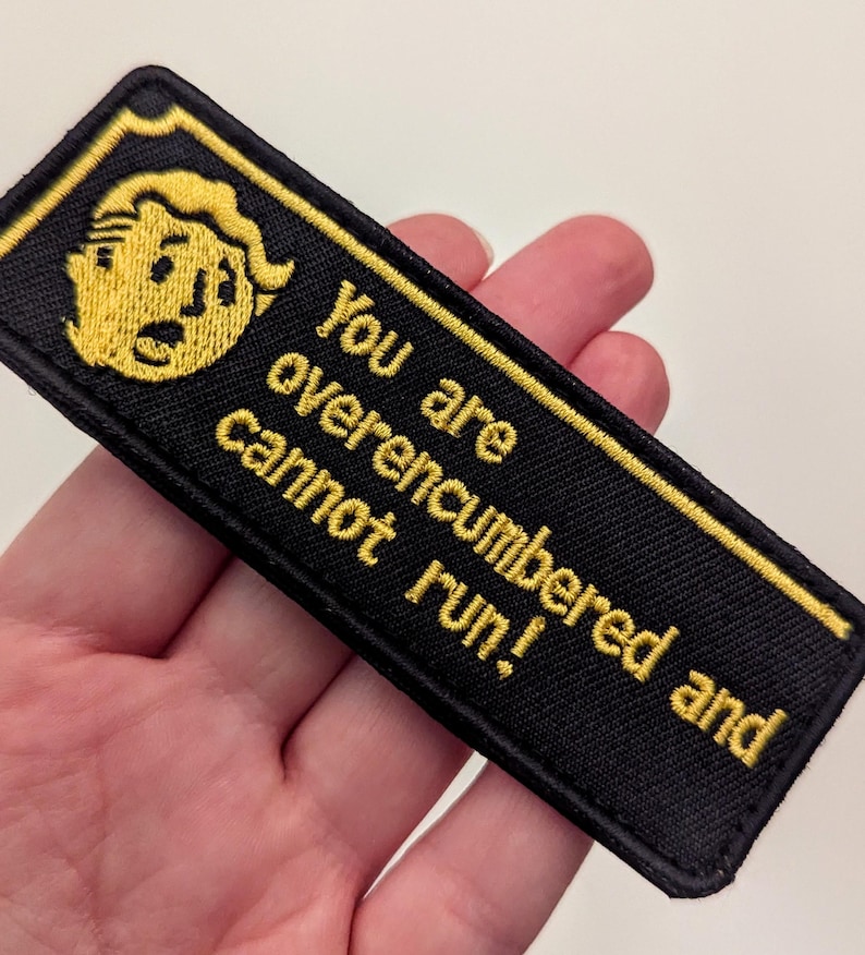 May include: Black rectangular embroidered patch with yellow text and a yellow cartoon character. The text reads "You are overencumbered and cannot run!" The patch has a yellow border.