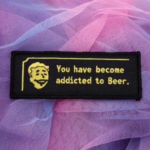 May include: A rectangular black embroidered patch featuring a yellow stitched graphic of a smiling character face on the left and the yellow embroidered text "You have become addicted to Beer." centred on the right side with a yellow border.