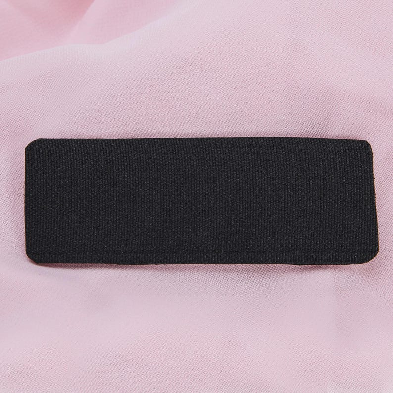 May include: A rectangular black fabric patch with rounded corners. The patch has a textured surface and is set against a soft, light pink background. The fabric appears to be tightly woven.
