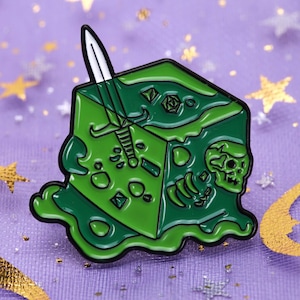 May include: Green enamel pin of a melting green d20 dice with a sword sticking out of it and a skull and bones inside.
