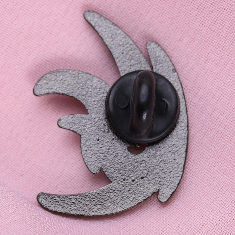 May include: A silver-colored enamel pin with a black clasp. The pin has a stylized, abstract design with curved shapes. The back of the pin is visible, showing the clasp and the textured surface of the metal.