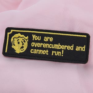 May include: Black rectangular patch with a yellow border and a yellow cartoon character on the left. The text "You are overencumbered and cannot run!" is embroidered in yellow.
