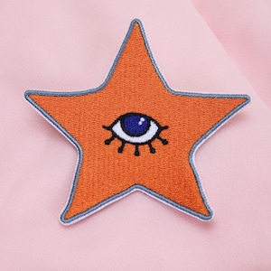 May include: Embroidered orange star patch with a blue eye design. The star has a light blue border and is set against a pink background. This patch is suitable for clothing or accessories.