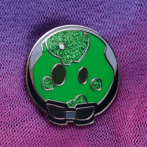 May include: A circular enamel pin with a green skull design and glitter detailing. The pin has a black border with gold trim and black sunglasses at the bottom. The background is a purple and pink gradient.