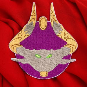Könnte beinhalten: Gestickter Aufnäher mit einem stilisierten Kreaturen-Design. Der Aufnäher hat einen violetten Mittelbereich mit einem beigen Gesicht und grünen Augen. Goldfarbene Hörner und Akzente umrahmen das Design, vor rotem Hintergrund.