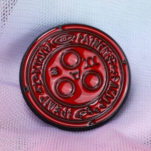 May include: A red and black enamel pin with a circular design. The centre of the pin features three circles with a symbol inside each circle. The outer circle has a black border and a repeating pattern of symbols.