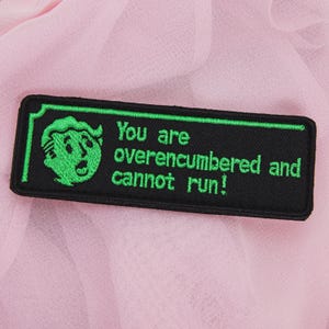 May include: Black rectangular patch with green border and text that reads "You are overencumbered and cannot run!" A green cartoon character is on the left side of the patch. The patch is on a pink fabric background.