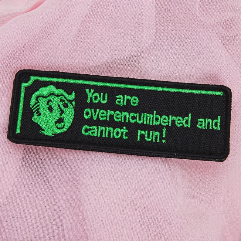 May include: Black rectangular patch with green border and text that reads "You are overencumbered and cannot run!" A green cartoon character is on the left side of the patch. The patch is on a pink fabric background.