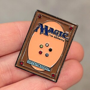 May include: A brown and black enamel pin featuring the Magic: The Gathering logo with a peach colored background and a red, green, white, black, and blue gem design. The pin says "Deckmaster" in a gold banner.