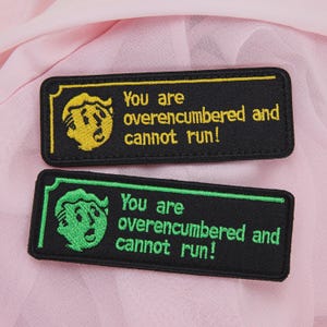 May include: Two rectangular embroidered patches with the text "You are overencumbered and cannot run!". One patch features a yellow design on a black background, the other has a green design. The patches are on a pink surface.