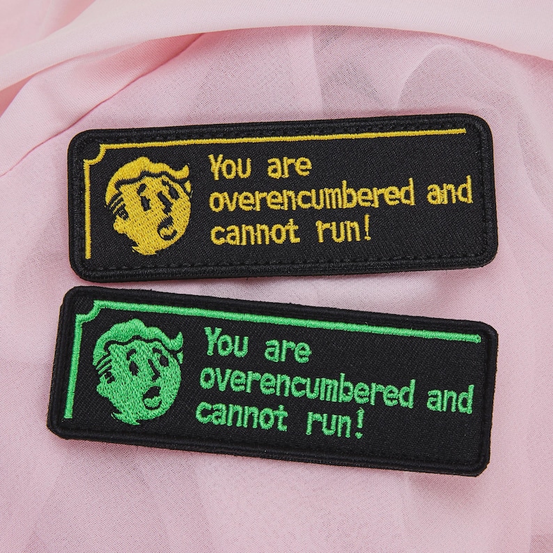 May include: Two rectangular embroidered patches with the text "You are overencumbered and cannot run!". One patch features a yellow design on a black background, the other has a green design. The patches are on a pink surface.