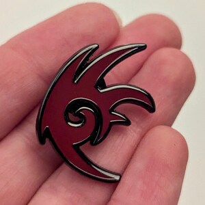 May include: A burgundy and black enamel pin featuring a stylized, abstract design. The pin has a glossy finish and a black outline, with a swirling detail in the center. The pin is held in a person's hand.