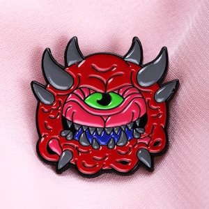 May include: A red enamel pin depicting a cartoon monster with a single green eye, grey horns, and sharp teeth. The monster's mouth is pink with blue detailing. The pin is set against a pink backdrop.