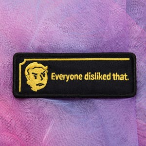 May include: Black rectangular patch with a yellow border and a yellow cartoon face on the left. The text "Everyone disliked that" is in yellow, centered on the patch. The patch is on a purple and pink background.