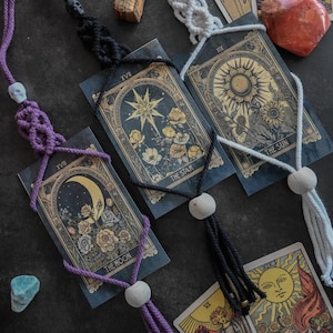 Könnte beinhalten: Draufsicht auf Tarotkarten mit Makramee-Lesezeichen. Die Karten zeigen himmlische und florale Illustrationen. Die Lesezeichen sind lila, schwarz und weiß, jedes mit einer Totenkopfperle und einer Quaste. Kristalle und andere Objekte sind um die Karten herum angeordnet.