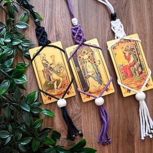 Könnte beinhalten: Drei Makramee-Wandbehänge mit Tarotkarten. Die linke Karte ist der Ritter der Kelche mit schwarzer Kordel, die mittlere die Hohepriesterin mit violetter Kordel und die rechte die Königin der Münzen mit weißer Kordel. Jede hat eine Totenkopfperle.