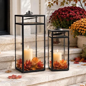 May include: Two rectangular black metal lanterns with clear glass panels, each containing a single cream-colored pillar candle surrounded by a base of vibrant orange, red, and yellow autumn maple leaves, positioned on stone steps.