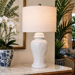 May include: A white ceramic table lamp with a textured wheat stalk pattern and a cylindrical fabric shade. The lamp features a smooth, glossy finish and a matching white lid-style top, standing on a base that tapers toward the center.