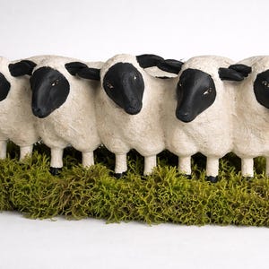 May include: A decorative sculpture featuring five sheep with black faces and ears, standing on a bed of green moss. The sheep are white with textured surfaces, creating a charming and rustic aesthetic.