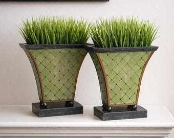 May include: Two decorative green planters with faux grass. The planters have a lattice design and are trimmed with brown and black accents. They sit on a black base with small ball feet. The planters are displayed on a white surface.