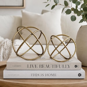 May include: Two antique brass-colored metal spherical sculptures with intersecting rings resting on two stacked books titled Live Beautifully and This Is Home. The spheres have a textured, metallic finish and are positioned on a wooden surface.