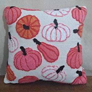 May include: A square throw pillow with a cream-colored fabric base featuring a tufted, textured pattern of pumpkins in various shades of pink, orange, and white. Each pumpkin has a small, dark brown stem detail embroidered onto the surface.