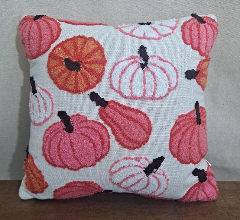May include: A square throw pillow with a cream-colored fabric base featuring a tufted, textured pattern of pumpkins in various shades of pink, orange, and white. Each pumpkin has a small, dark brown stem detail embroidered onto the surface.