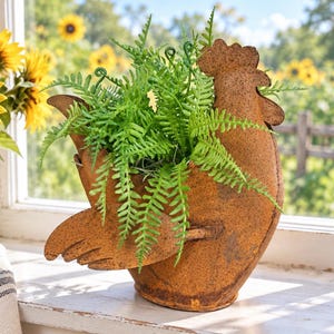 May include: A rooster-shaped metal planter with a weathered, rusted brown finish. The hollow body of the rooster serves as a container for a vibrant green artificial fern plant with delicate, feathery fronds that spill over the sides.