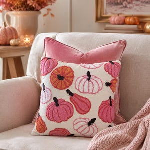 May include: A square throw pillow featuring a cream-colored fabric base with a repeating pattern of embroidered pumpkins in shades of pink, orange, and red. The pillow has a textured, tufted surface and is layered in front of a solid pink velvet pillow.