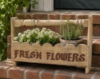 May include: A rustic wooden flower box with a handle, filled with white flowers and a potted herb. The box is painted with the words "FRESH FLOWERS" in brown. The box is sitting on a brick surface.