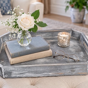 May include: A gray, octagonal wooden tray with a distressed finish and cutout handles. The tray holds a clear glass vase with a white rose and baby's breath, two stacked hardcover books, a small metallic candle holder, and round glasses.
