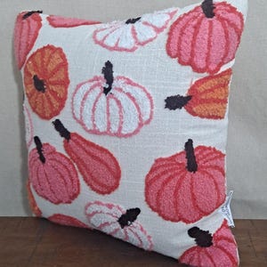May include: A square throw pillow with a cream-colored fabric base featuring a tufted, textured pattern of pumpkins in shades of pink, orange, and white. Each pumpkin has a dark brown stem. A small white fabric tag is visible on the lower right side.