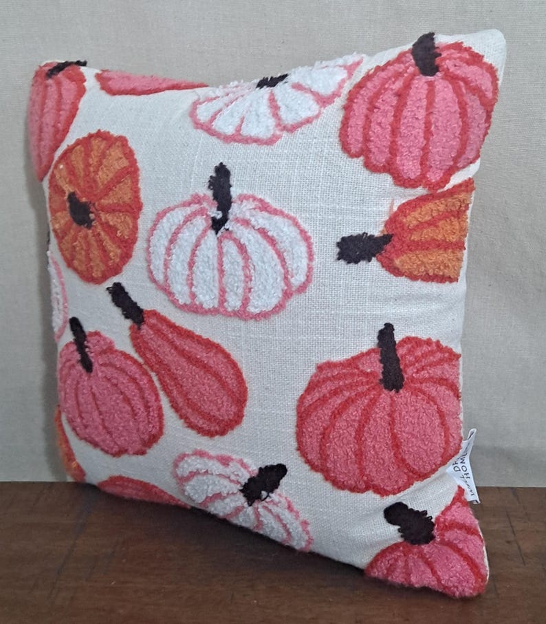 May include: A square throw pillow with a cream-colored fabric base featuring a tufted, textured pattern of pumpkins in shades of pink, orange, and white. Each pumpkin has a dark brown stem. A small white fabric tag is visible on the lower right side.