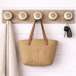 May include: A wooden wall-mounted coat rack with five decorative knobs. A beige towel, a woven tote bag with brown leather handles, and a set of keys hang from the hooks. The knobs have a cream and gold floral design.